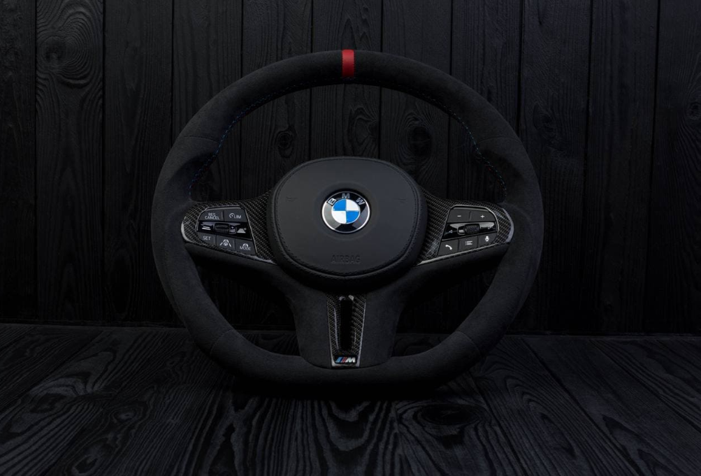 A black BMW steering wheel, showing control buttons on both sides and a central logo, is set against a dark wood-paneled background. Text includes: "BMW," "AIRBAG," and various button labels.