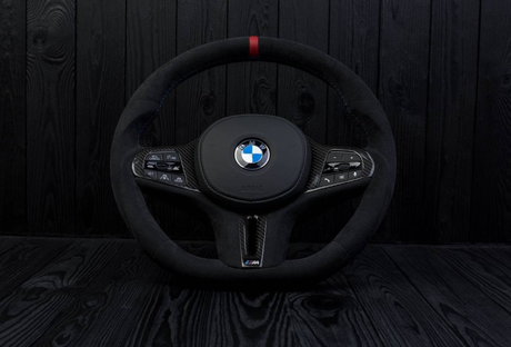 A black BMW steering wheel, showing control buttons on both sides and a central logo, is set against a dark wood-paneled background. Text includes: "BMW," "AIRBAG," and various button labels.
