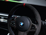 A steering wheel with carbon fiber accents and colored stitching is mounted in a car interior. The dashboard features a digital display and other controls, with the BMW logo at the center.