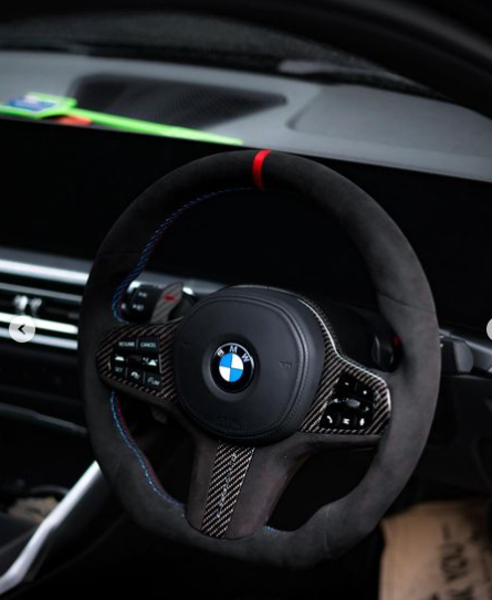 A steering wheel with carbon fiber accents and colored stitching is mounted in a car interior. The dashboard features a digital display and other controls, with the BMW logo at the center.
