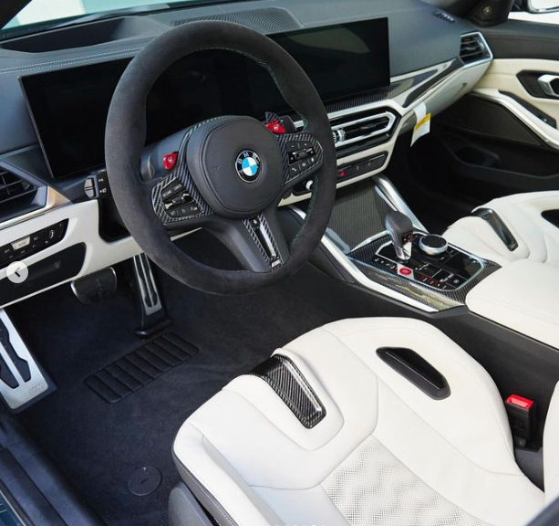 Steering wheel displays BMW logo, set in a modern car interior with dual digital screens, white leather seats, and a central console featuring a gear shifter and control knobs.