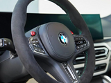 A sleek steering wheel adorned with a BMW logo features black leather and carbon fiber details. It includes control buttons and sits within a modern car interior with digital displays.