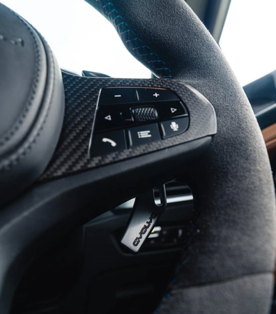 Steering wheel featuring textured grip, control buttons for phone, voice, and volume on carbon-fiber panel. Blue stitching detail, 'Evolve' tag visible, inside a car interior.