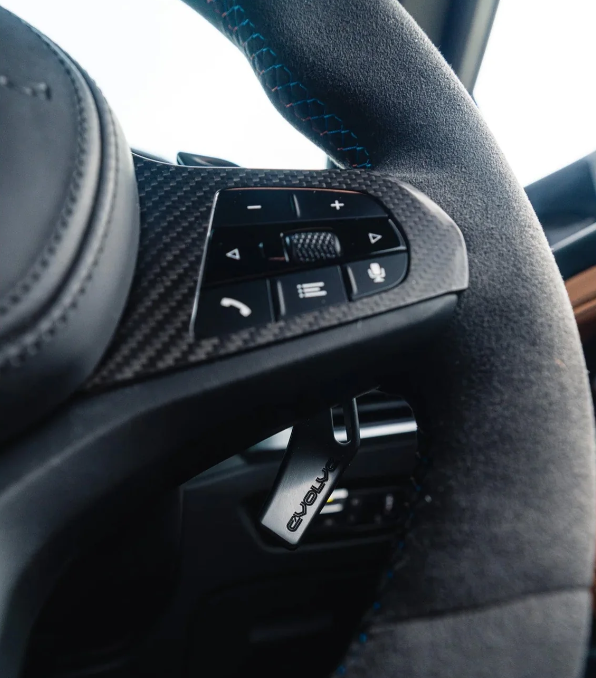 Steering wheel featuring textured grip, control buttons for phone, voice, and volume on carbon-fiber panel. Blue stitching detail, 'Evolve' tag visible, inside a car interior.