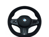 A round, black BMW steering wheel, featuring control buttons, is set against a plain white background. BMW logo in the center, 