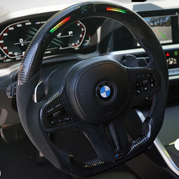 A carbon fiber BMW steering wheel features illuminated indicators. Buttons for cruise control and audio settings are integrated. It's set within a sleek car dashboard, displaying speed and navigation information.