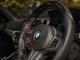 The carbon fiber steering wheel of a BMW is stationary, surrounded by a sleek car interior with visible controls and vents. Sunlit trees are seen through the window.