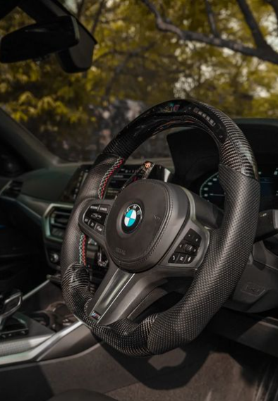 The carbon fiber steering wheel of a BMW is stationary, surrounded by a sleek car interior with visible controls and vents. Sunlit trees are seen through the window.