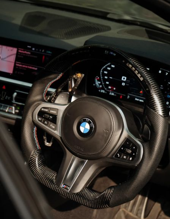 A BMW steering wheel with carbon fiber detail is centered, featuring control buttons. Behind it, a dashboard displays digital gauges and a navigation screen in a modern car interior.