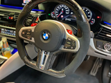A BMW steering wheel rests in a car's interior. Dashboard displays digital gauges, and an infotainment screen shows music controls. Various buttons and controls are visible on the console.