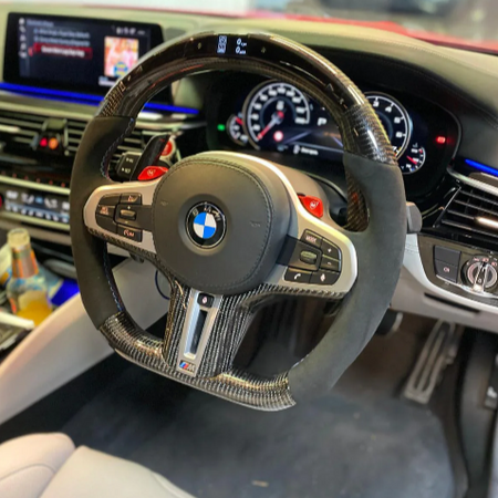 A BMW steering wheel rests in a car's interior. Dashboard displays digital gauges, and an infotainment screen shows music controls. Various buttons and controls are visible on the console.