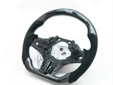 A steering wheel with a partial carbon fiber finish and exposed inner mechanism rests on a white surface, showing blue and red wires extending from its left side.