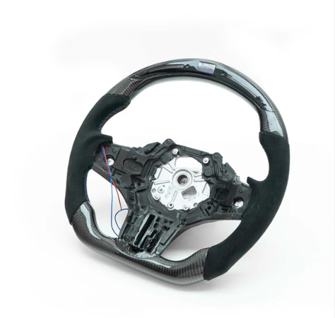 A steering wheel with a partial carbon fiber finish and exposed inner mechanism rests on a white surface, showing blue and red wires extending from its left side.