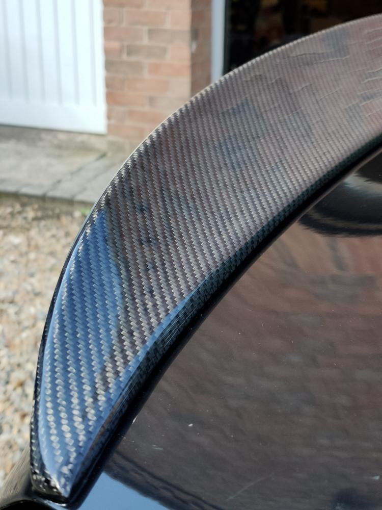 A carbon fiber car spoiler rests on a glossy black surface, outdoors near a brick wall and gravel pathway, with a white garage door in the background.