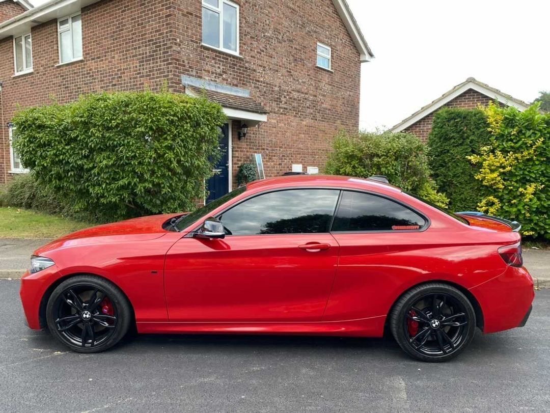 A red sports car is parked on a residential street, with a brick house and green bushes in the background. The car features tinted windows and black wheels.