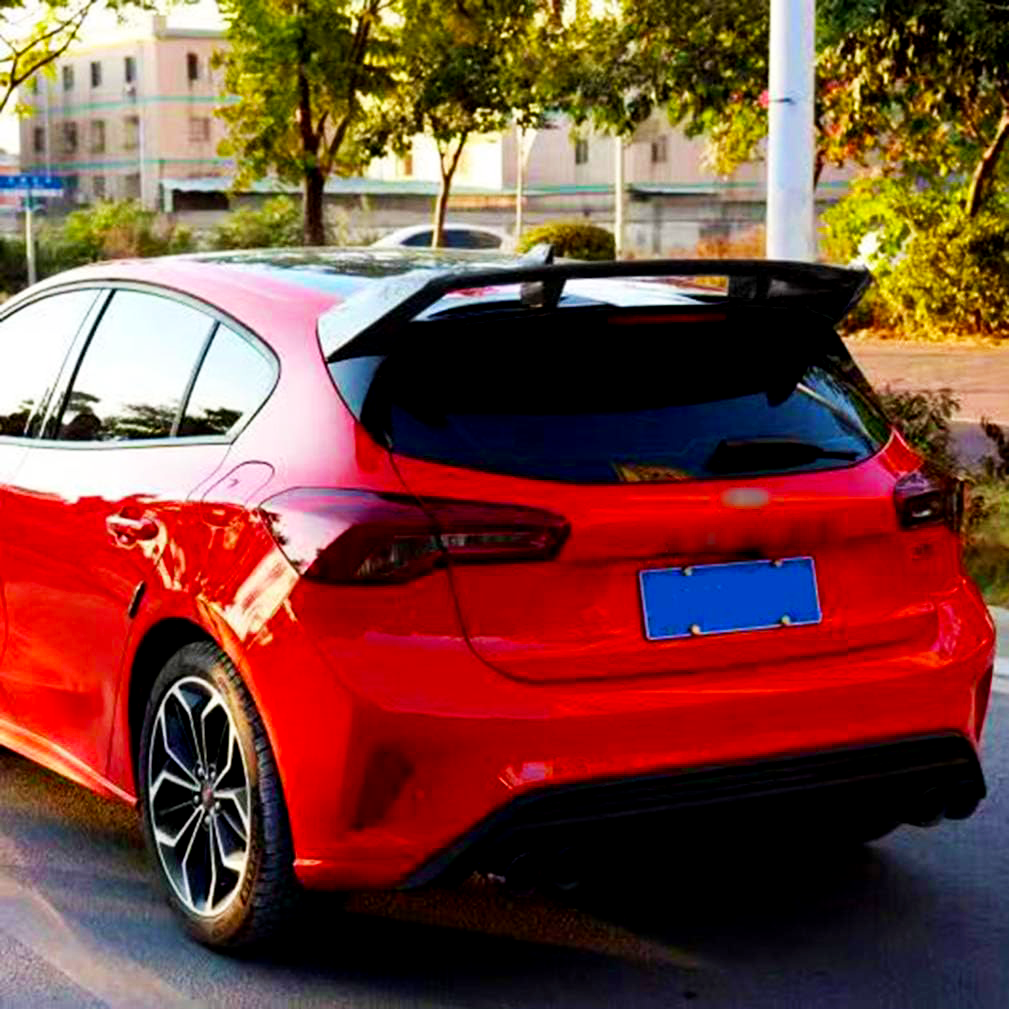 A red car is parked on a street; its sleek design features a sporty rear spoiler. The surrounding area is leafy with residential buildings in the background. The license plate is blue.