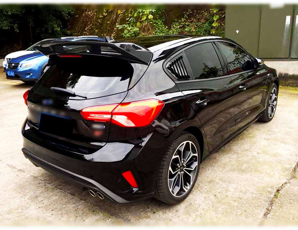 A sleek black hatchback car is parked on a concrete lot, surrounded by greenery and other vehicles. It features tinted windows, stylish taillights, and dual exhaust pipes.