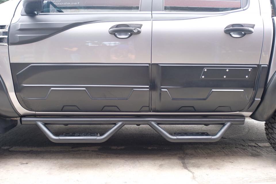 A rugged vehicle features side protective bars and reinforced step plates along two tightly sealed, dark-colored doors, parked on a concrete surface with large off-road tires partially visible.