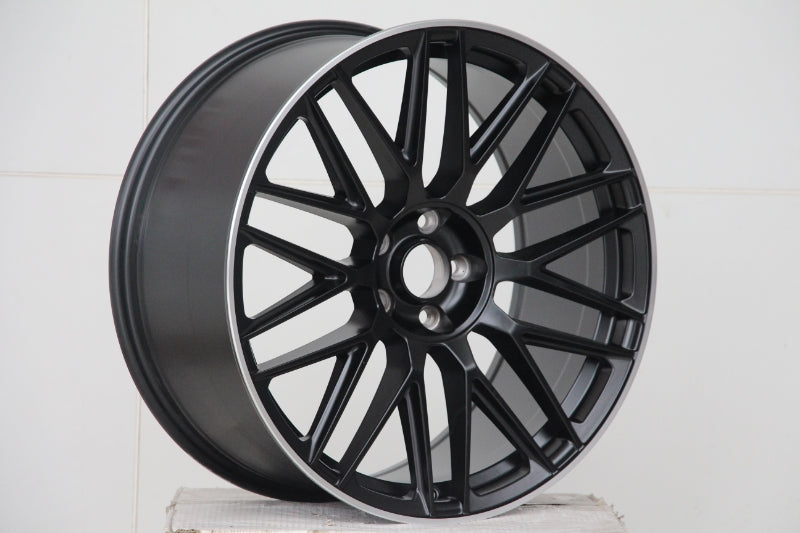A black alloy car wheel with intricate spoke design rests on a plain white surface against a tiled white background, emphasizing the wheel's sleek, modern appearance.