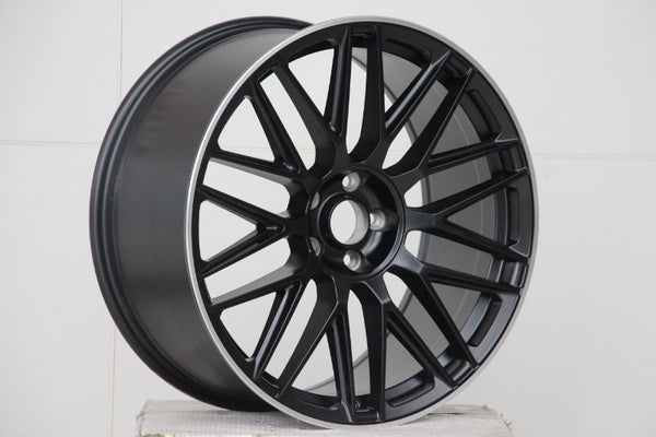 A black alloy car wheel with intricate spoke design rests on a plain white surface against a tiled white background, emphasizing the wheel's sleek, modern appearance.