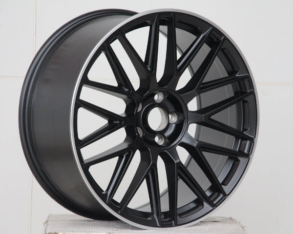 A matte black alloy wheel with a multi-spoke design is resting on a white, textured surface, set against a plain white background.
