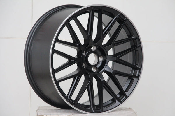 A matte black alloy wheel with a multi-spoke design is resting on a white, textured surface, set against a plain white background.