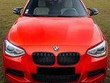 A bright orange car, stationary, is parked on a grassy field. It features a distinctive grille with a BMW emblem and sleek headlights, surrounded by an open area with distant buildings.
