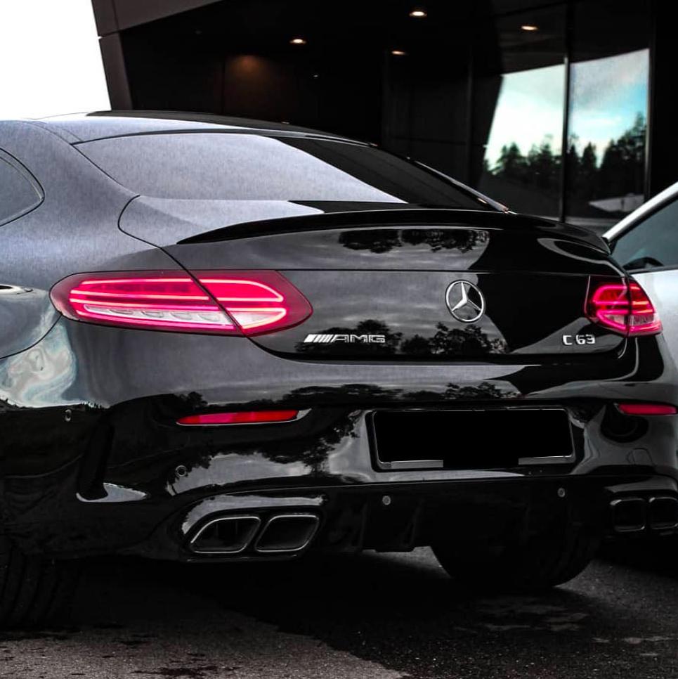 A black Mercedes-Benz AMG C63 parked, with illuminated rear lights, chrome quad exhausts, and badges. It is set against a modern building with reflective windows and dim lighting.