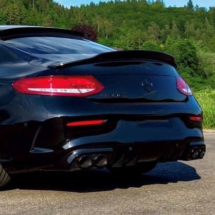 A sleek black car with dual exhausts is parked on a road. The background features lush greenery and trees under a clear blue sky.