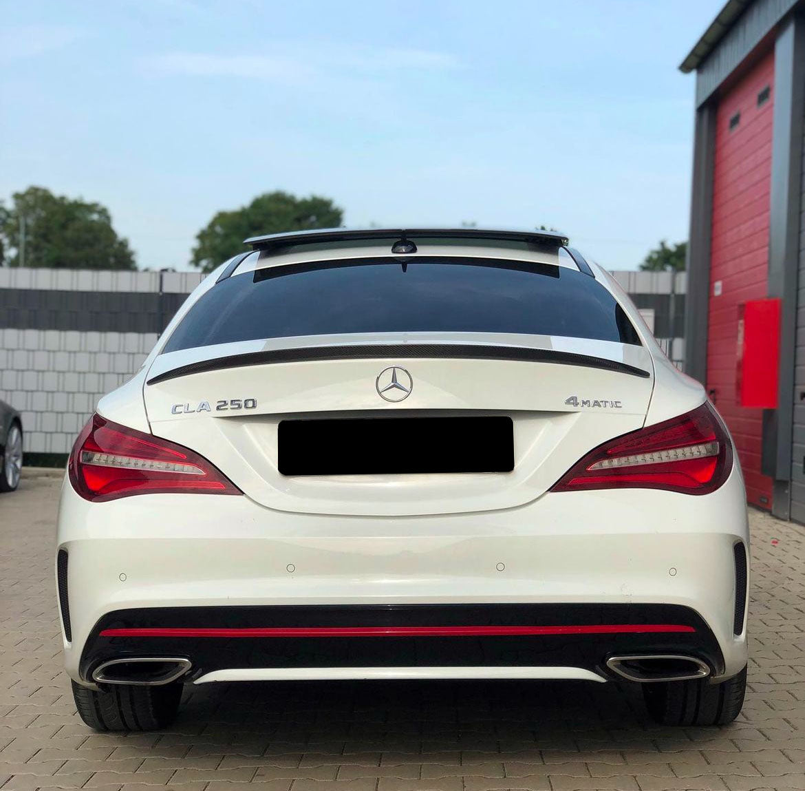 A white Mercedes-Benz CLA 250 with "4MATIC" badge is parked in front of a garage with red doors and gray walls. The background includes trees and another vehicle.