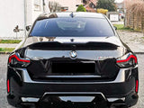 A black BMW M240i is parked on a driveway, surrounded by residential houses and green lawn. The car's prominent features include taillights and visible badges reading 