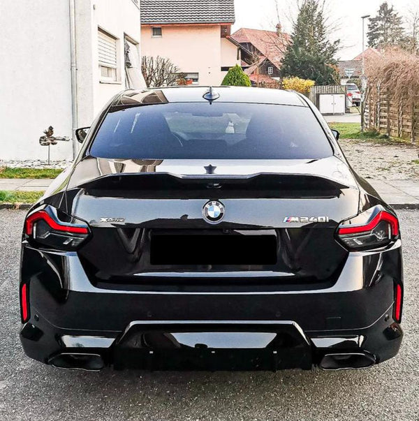 A black BMW M240i is parked on a driveway, surrounded by residential houses and green lawn. The car's prominent features include taillights and visible badges reading "X Drive" and "M240i".