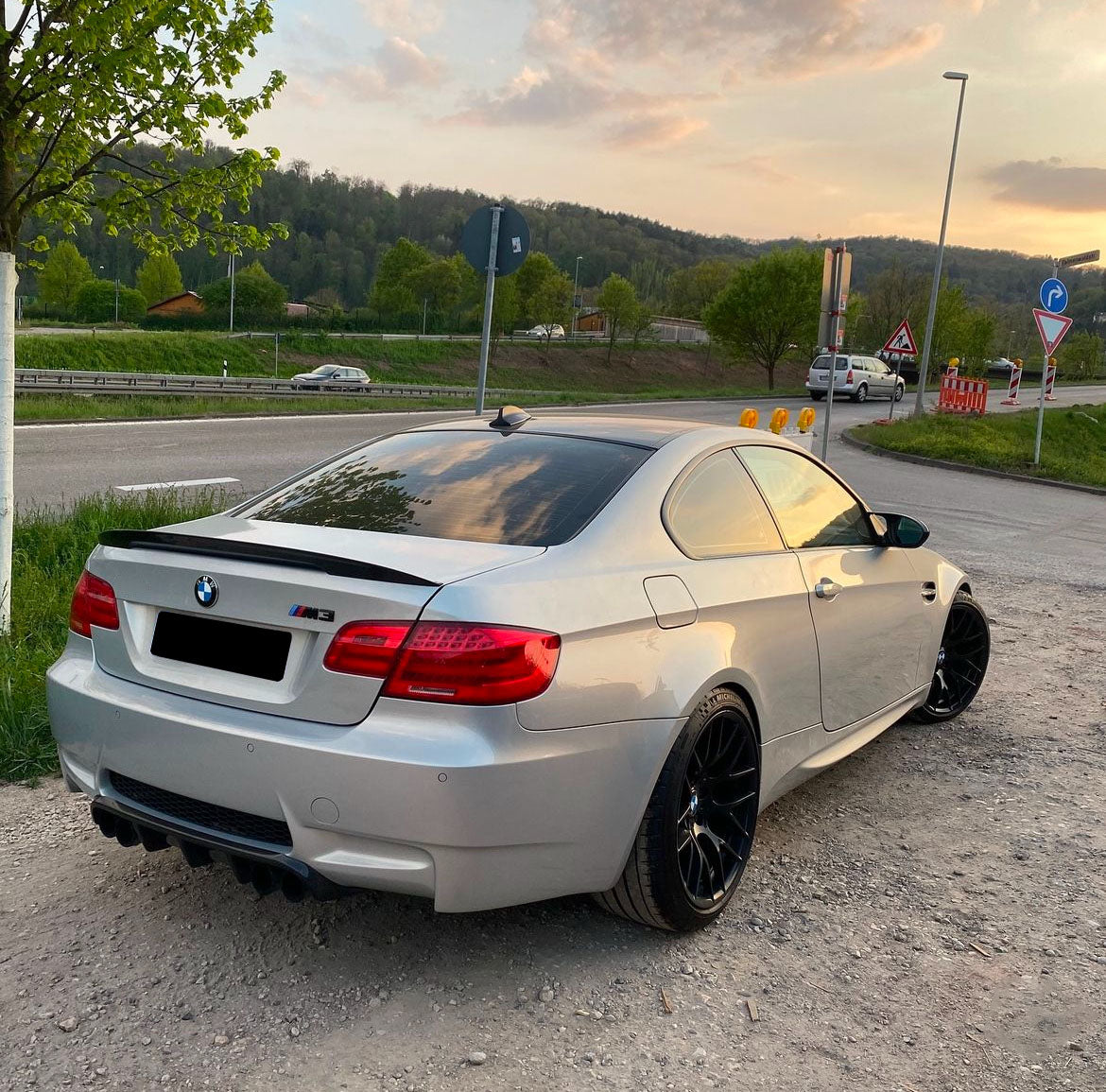 A silver BMW M3 is parked on a gravel area beside a road, with a backdrop of greenery and trees under a partly cloudy sunset sky.