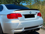 A white BMW M3 is parked on the street, surrounded by tall grass and trees. The car's rear view showcases its taillights and four exhaust pipes.