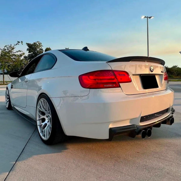 A white sports car with sleek, aerodynamic design is parked on a concrete surface, featuring dual exhausts and large alloy wheels, under clear blue sky with scattered greenery in the background.