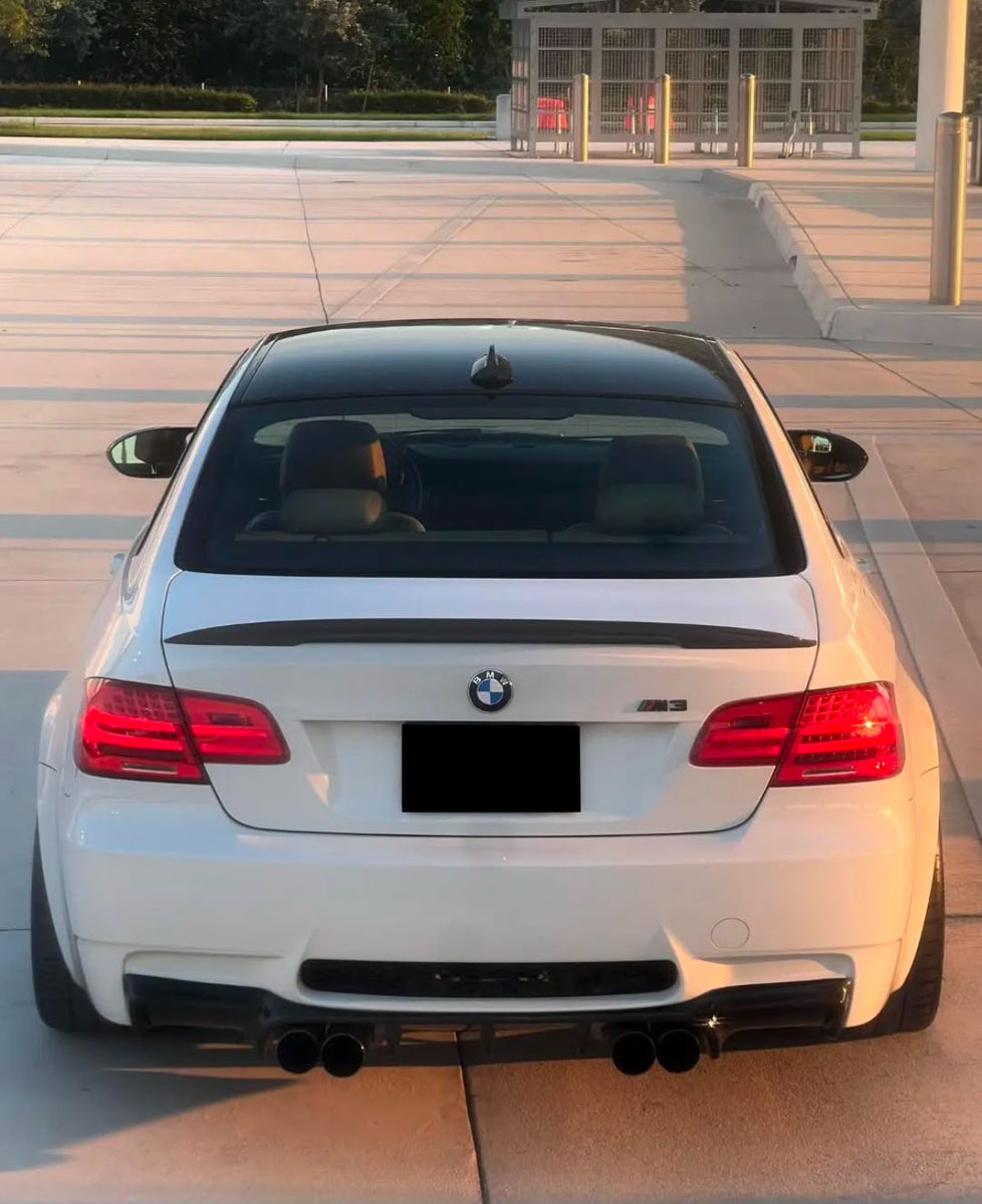 A white BMW M3 is parked on a sunlit pavement. The rear view shows its dual exhausts, taillights, and the BMW emblem. A modern building with poles is visible in the background.