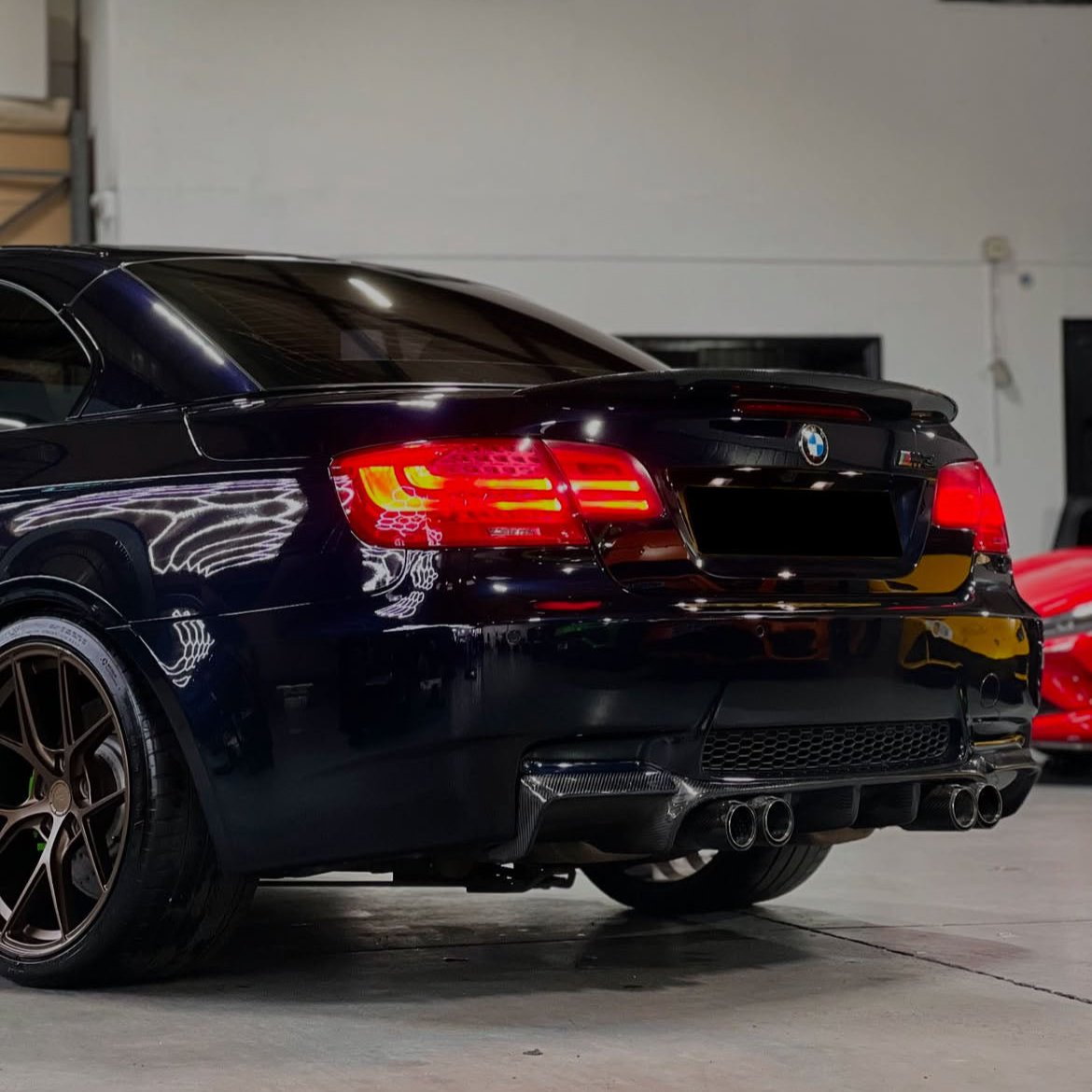 A black car with illuminated rear lights and dual exhaust system is parked in a garage with a red vehicle in the background. The environment is well-lit.