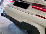 A white BMW 330i has its rear view with illuminated taillights and visible exhaust pipes. It's parked indoors, near a framed picture and red-black furniture.