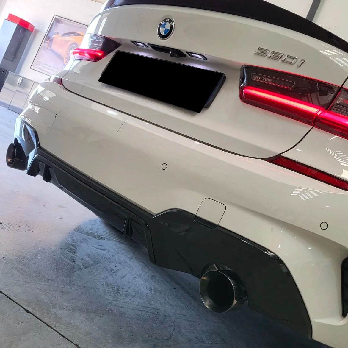 A white BMW 330i has its rear view with illuminated taillights and visible exhaust pipes. It's parked indoors, near a framed picture and red-black furniture.