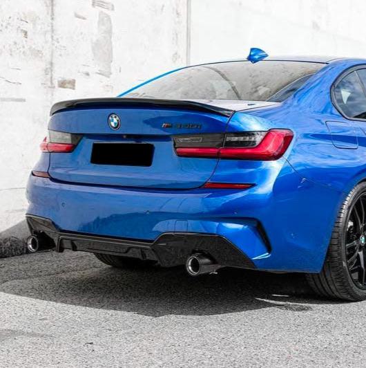 A blue BMW car is parked on a street, against a white wall. The model is identified as "M340i," with black rear license plate and twin exhaust pipes.