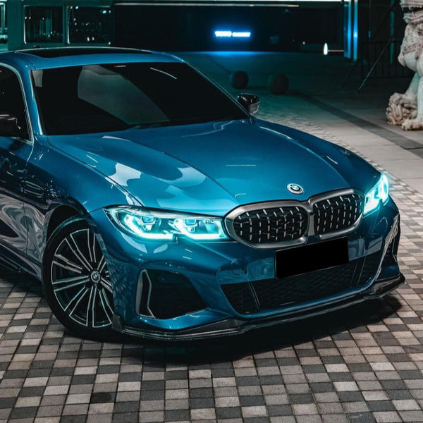 A blue car with illuminated headlights is parked on a checkered pavement beside a building and a statue, under ambient lighting.