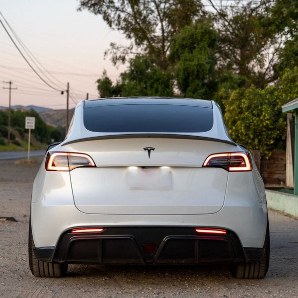 A white sedan with a lit rear logo and taillights is parked on a gravel road. Trees and power lines are visible under the evening sky in the background.