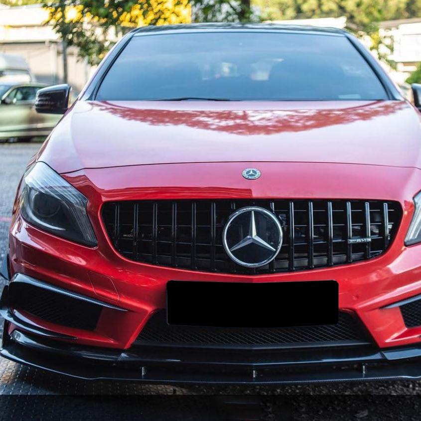 A red Mercedes-Benz car, viewed from the front, is parked. It features a prominent grille with the Mercedes emblem. Other cars and greenery are visible in the background.