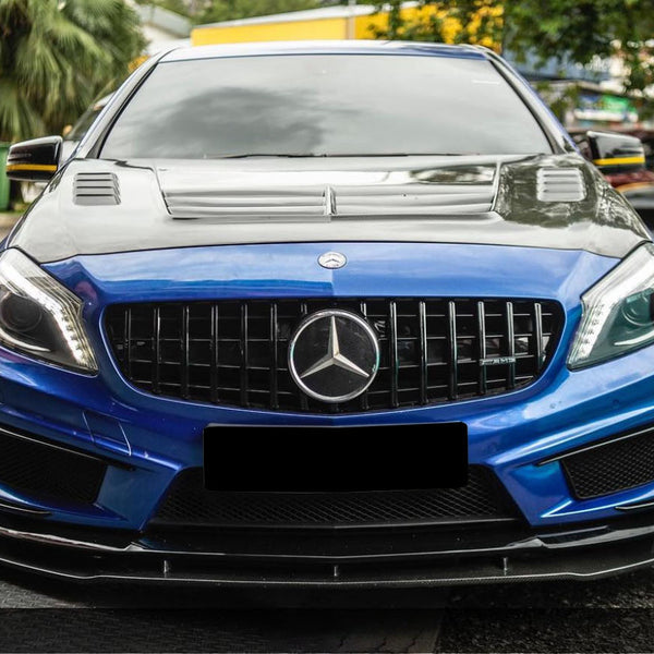 A blue Mercedes-Benz car is parked on a street, featuring a blacked-out grille and hood with vents. Surrounding it are trees and other parked vehicles in an outdoor setting.