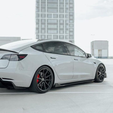 A white car with dark rims is parked on a rooftop parking lot, with a tall building visible in the background.
