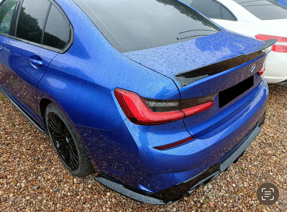 Blue car parked on gravel, covered in rain droplets, adjacent to a white car. The rear view highlights sleek, angular tail lights and a black rear spoiler.