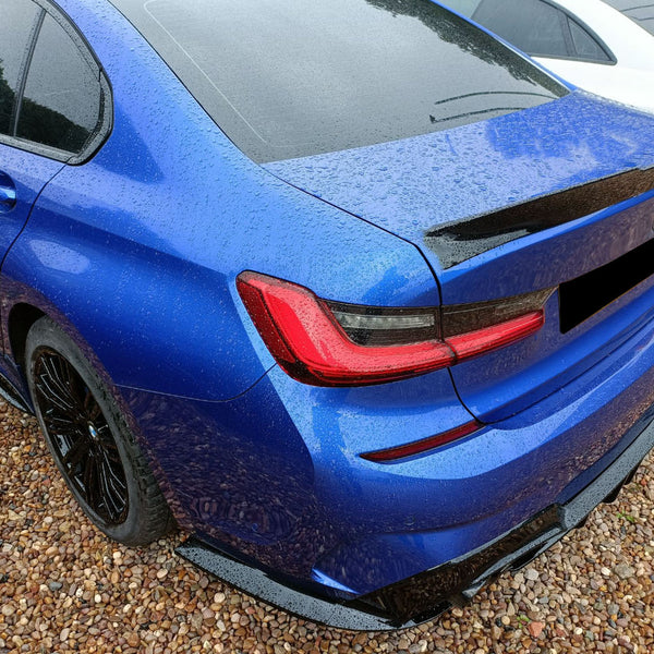 Blue car parked on gravel, covered in rain droplets, adjacent to a white car. The rear view highlights sleek, angular tail lights and a black rear spoiler.
