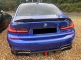 A blue BMW car is parked on a gravel surface, displaying a sleek rear with dual exhausts. It sits beside another vehicle, surrounded by lush greenery.
