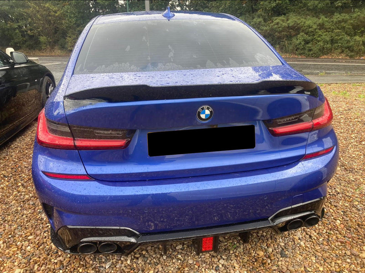A blue BMW car is parked on a gravel surface, displaying a sleek rear with dual exhausts. It sits beside another vehicle, surrounded by lush greenery.