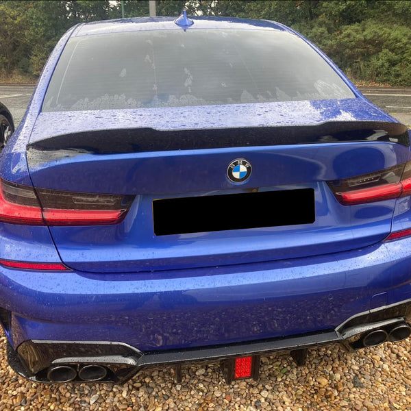 A blue BMW car is parked on a gravel surface, displaying a sleek rear with dual exhausts. It sits beside another vehicle, surrounded by lush greenery.